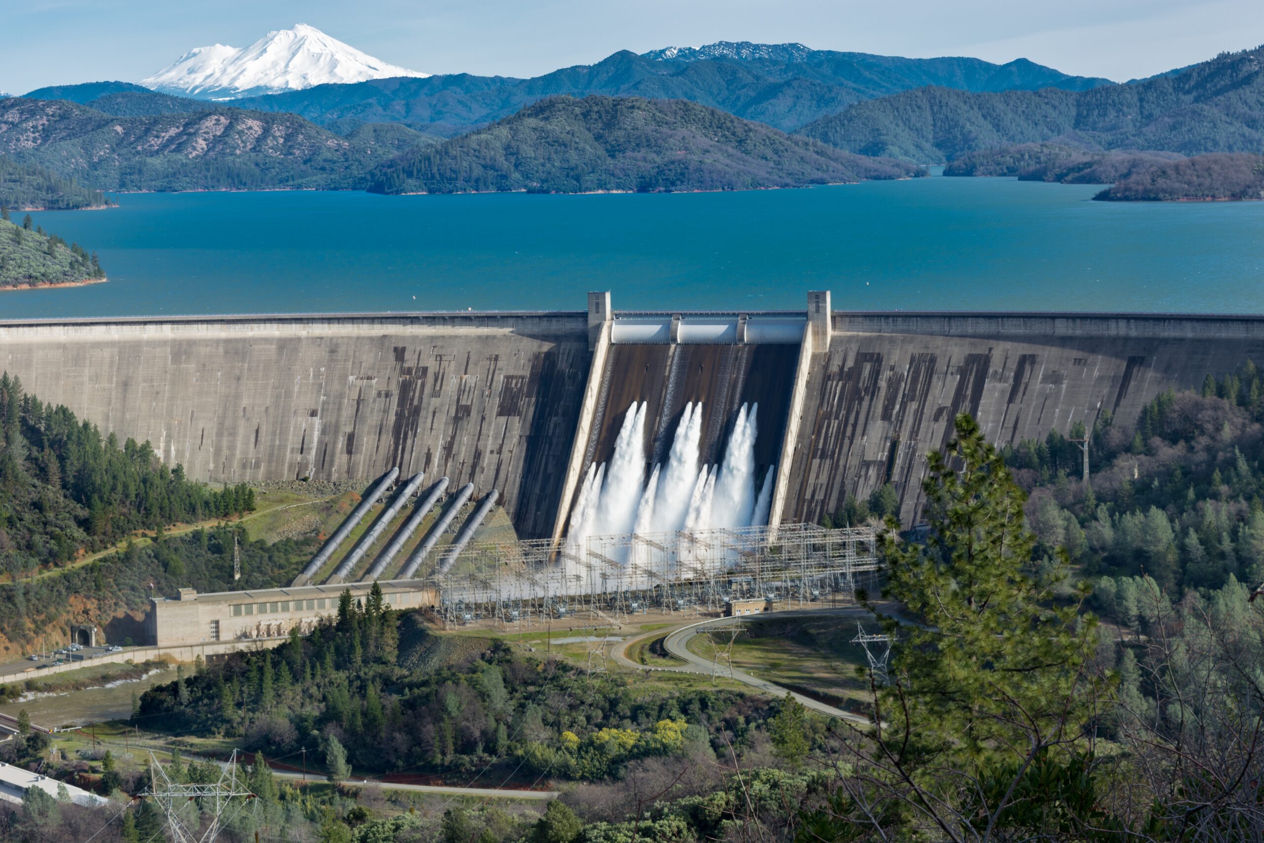 Picture of Shasta Dam surrounded by roads and trees with a lake and mountains on the background